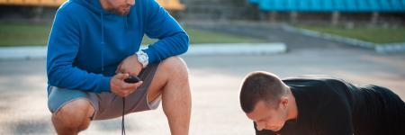a trainer is crouched down, timing a man doing a plank