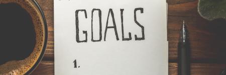a notepad with the word "goals" written on it sits on a wooden table next to a coffee mug and pen
